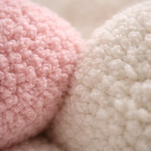 May include: Close-up of a soft, textured fabric in two colors: light pink and off-white. The material has a fluffy, looped pile, creating a cozy and inviting appearance. The image highlights the tactile quality of the textile.