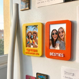 May include: Two rectangular photo frames, one yellow and one orange, are attached to a white surface. The yellow frame has the word "SPAIN" and "SUMMER 23" in relief. The orange frame has the word "BESTIES" in relief. Each frame holds a photo of smiling people.