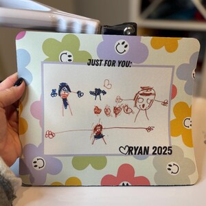May include: A rectangular mousepad with a floral pattern in pastel colors. The center features a child's drawing with the text "JUST FOR YOU:" and "RYAN 2025". The mousepad is held by a hand.