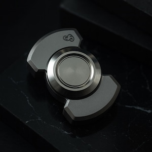 Puede incluir: Un spinner plateado con un componente central circular y tres brazos curvos. El spinner tiene un acabado de metal cepillado y un pequeño emblema en un brazo. El fondo es una superficie oscura y texturizada.