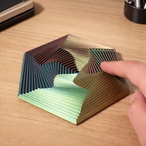 Op de afbeelding: Een veelkleurig, geometrisch, zeshoekig object met een spiraalvormig ontwerp. Het object heeft tinten groen, bruin en blauw. Een vinger raakt het oppervlak van het object. Het object staat op een houten oppervlak.
