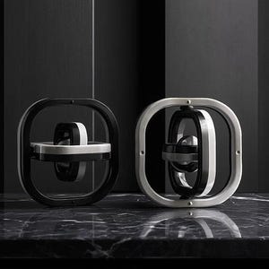 以下が含まれることがあります： モダンな幾何学的な彫刻が2つ。1つは白のアクセントが付いた黒、もう1つは黒のアクセントが付いたシルバーです。それぞれ正方形の外枠と回転する内側の構造を持っています。彫刻は暗く、質感のある表面に置かれています。