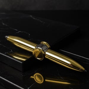 May include: A gold-colored pen with a silver band in the center, resting on a black marble surface. The pen's design is sleek and modern, with a polished finish. The pen is a writing instrument.