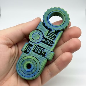Könnte beinhalten: Ein blau-grünes, 3D-gedrucktes Fidget-Spielzeug. Das Design ist komplex und weist kreisförmige und geometrische Elemente auf. Das Spielzeug wird in einer Hand gehalten, vor einem weißen Hintergrund.