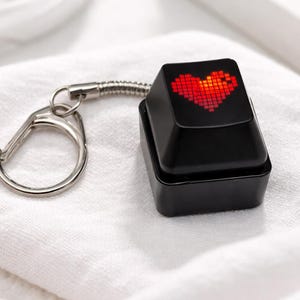 Puede incluir: Llavero negro con un diseño de corazón rojo pixelado sobre un cuadrado negro. El llavero tiene un cierre plateado y un conector metálico flexible. El artículo está sobre una superficie blanca texturizada.
