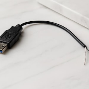 Puede incluir: Un cable USB negro con un conector USB en un extremo y cables expuestos en el otro. El conector USB tiene un interior azul y un símbolo USB. El cable está sobre una superficie de mármol blanco.