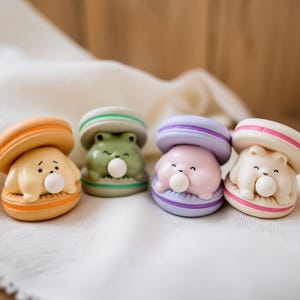 Puede incluir: Cuatro figuras coloridas con forma de macaron. Cada una tiene un personaje animal diferente: un perro, una rana, una criatura rosa y un oso. Los macarons son naranjas, verdes, morados y rosas, con detalles blancos y pastel. Cada figura tiene una burbuja.