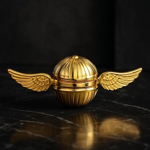 May include: A golden sphere with two wings, resembling a Snitch. The sphere has a ribbed design and a small knob on top. The wings are detailed with feather patterns. The object is set against a dark background.
