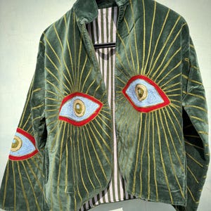 Könnte beinhalten: Dunkelgrüne Samtjacke mit goldgestickten Sonnenstrahlenmustern, die von großen Augenapplikationen ausgehen. Die Augen sind blau mit roten Umrissen und goldenen Details. Die Jacke hat ein gestreiftes Futter und wird auf einem schwarzen Kleiderbügel präsentiert.