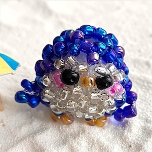 May include: A small, handcrafted beaded penguin figurine constructed from iridescent blue, silver, and black glass beads. The penguin features two black bead eyes, a small yellow beak, and is positioned next to a miniature yellow and blue beach umbrella.