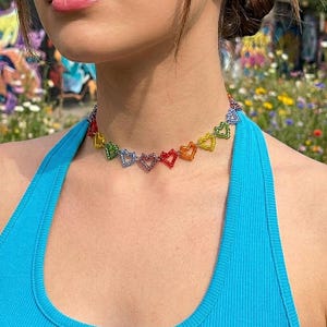 May include: A choker necklace featuring a series of small, connected heart-shaped charms in a rainbow spectrum of colors including red, orange, yellow, green, blue, and purple. The hearts are linked together to form a continuous chain around the neck.