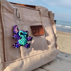 Handmade purple and blue beaded Stitch keychain hanging on a beige canvas tote bag. Scenic beach background with ocean waves and soft sunlight, showcasing the character charm as a stylish bag accessory.