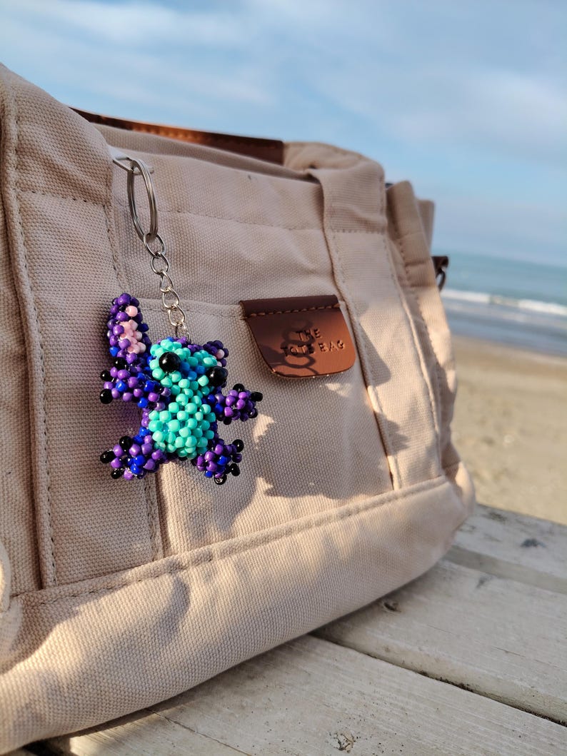 Handmade purple and blue beaded Stitch keychain hanging on a beige canvas tote bag. Scenic beach background with ocean waves and soft sunlight, showcasing the character charm as a stylish bag accessory.