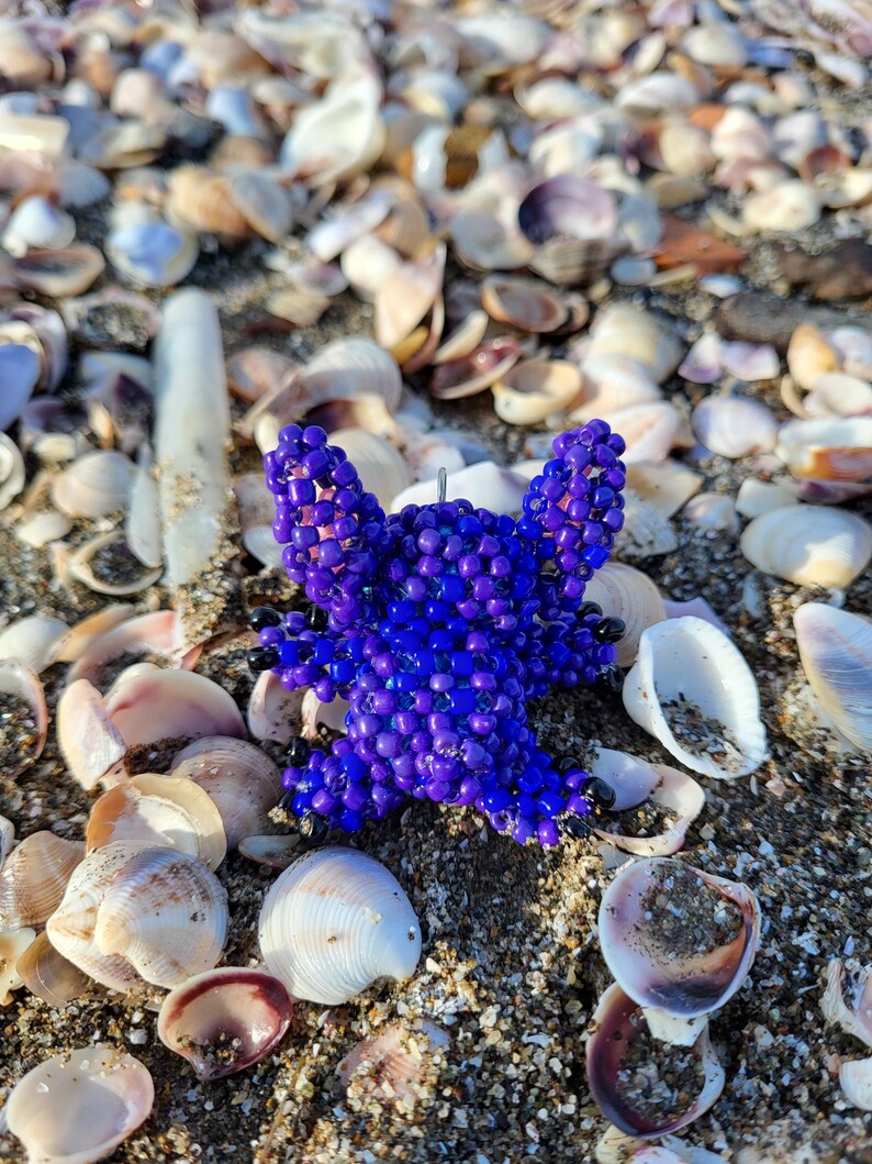 Pu&ograve; includere: Una stella marina artigianale di perline blu scuro poggia su una spiaggia sabbiosa cosparsa di conchiglie. La stella marina &egrave; fatta di piccole perline rotonde e ha cinque braccia. Lo sfondo presenta varie conchiglie nei toni del bianco, marrone e beige.
