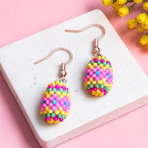 Puede incluir: Un par de pendientes de cuentas coloridas. Cada pendiente presenta un diseño en forma de lágrima, cubierto de pequeñas cuentas en tonos rosa, amarillo, verde y azul. Los pendientes cuelgan de un gancho simple.