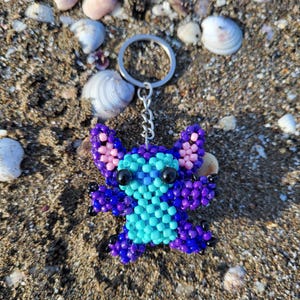 Handmade beaded Stitch keychain in vibrant purple and turquoise blue, lying on a sandy beach with natural sea shells. Detailed 3D beadwork character charm for bags or keys, unique gift for Disney and Lilo & Stitch fans.