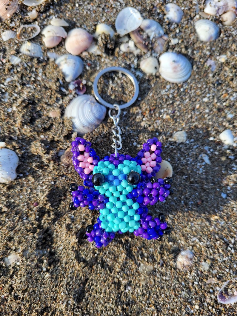 Handmade beaded Stitch keychain in vibrant purple and turquoise blue, lying on a sandy beach with natural sea shells. Detailed 3D beadwork character charm for bags or keys, unique gift for Disney and Lilo & Stitch fans.