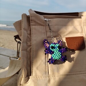 Purple beaded Stitch character keychain attached to a neutral color bag. Outdoor beach setting, handmade jewelry accessory for Lilo and Stitch lovers.
