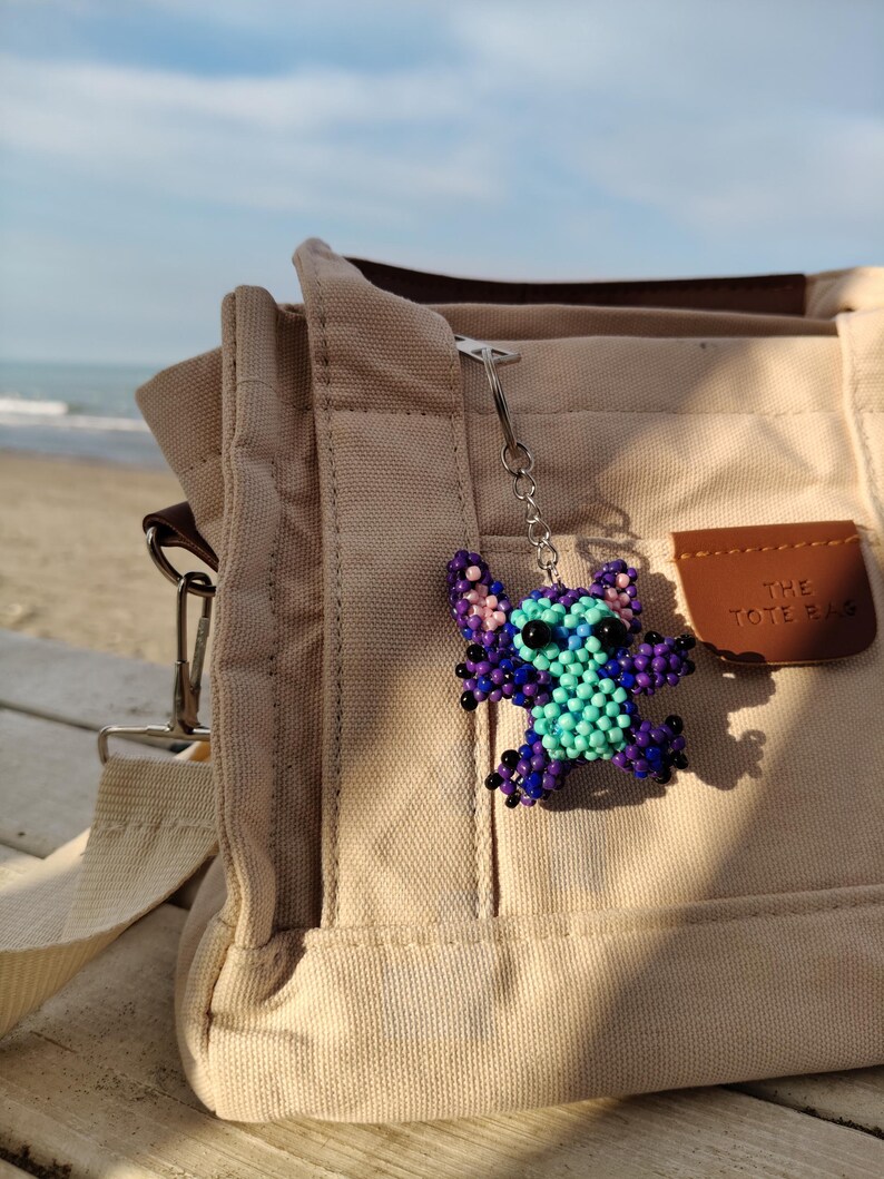 Purple beaded Stitch character keychain attached to a neutral color bag. Outdoor beach setting, handmade jewelry accessory for Lilo and Stitch lovers.