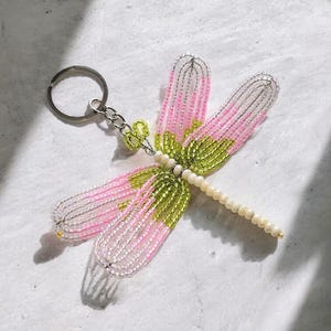 May include: A dragonfly keychain crafted from small beads. The wings feature pink and silver glass beads with lime green accents near the body. The body is composed of a row of cream-colored beads, and a silver metal ring is attached to the head.