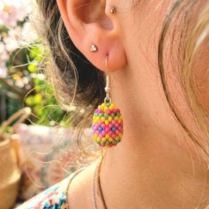 May include: Close-up of a colorful beaded earring. The earring is a vibrant mix of yellow, pink, blue, and green beads, suspended from a silver hook. The earring's design is oval-shaped, showcasing a playful and artistic style.