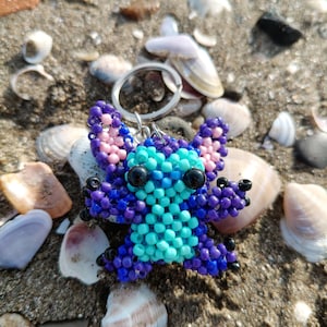 Close-up of a handcrafted beaded Stitch figurine keychain lying on a sandy beach surrounded by colorful sea shells. Detailed 3D bead art in vibrant purple and bright blue.