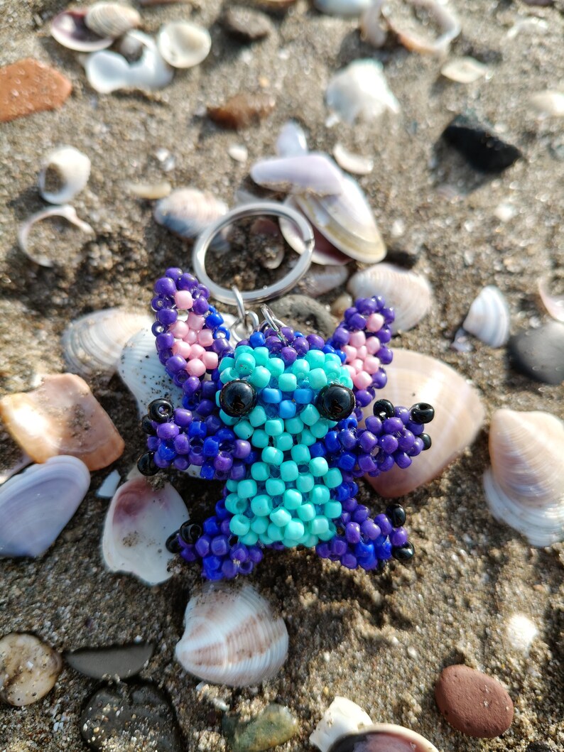 Close-up of a handcrafted beaded Stitch figurine keychain lying on a sandy beach surrounded by colorful sea shells. Detailed 3D bead art in vibrant purple and bright blue.