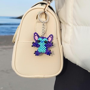 Pu&ograve; includere: Un portachiavi di perline raffigurante un personaggio con perline blu, viola e rosa. Il portachiavi &egrave; attaccato a una borsa beige con un anello e una catena di metallo color oro.