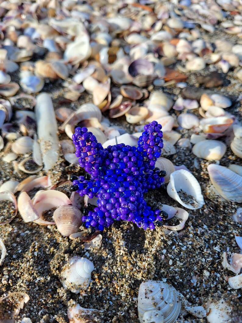 Close-up of a handcrafted beaded Stitch figurine keychain lying on a sandy beach with natural sea shells. Detailed 3D beadwork in vibrant purple and bright blue, perfect for animation fans and collectors.
