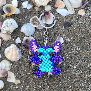 Handmade beaded Stitch keychain in vibrant purple and turquoise blue, lying on a sandy beach with natural sea shells. Detailed 3D beadwork character charm for bags or keys, unique gift for Disney and Lilo & Stitch fans.