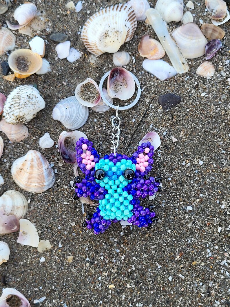 Handmade beaded Stitch keychain in vibrant purple and turquoise blue, lying on a sandy beach with natural sea shells. Detailed 3D beadwork character charm for bags or keys, unique gift for Disney and Lilo & Stitch fans.