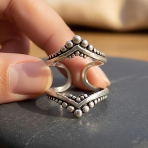 May include: Two silver rings with a beaded design are held up against a dark gray surface. The rings have a V-shape with a row of small beads along the top and bottom edges. The rings are connected by a curved band.