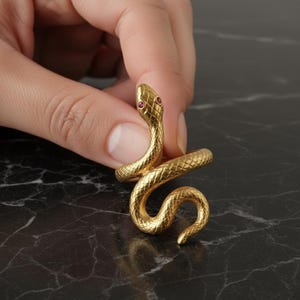 May include: A gold-colored snake ring with red gemstone eyes. The ring is coiled in a spiral shape, with detailed scales and a textured surface. The snake's head is raised, creating a striking and elegant jewelry piece.