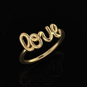 以下が含まれることがあります： 筆記体で「love」と書かれたゴールドカラーのリング。リングは光沢のある反射性の金属でできており、黒い背景に配置されています。「love」の文字が主なデザイン要素です。