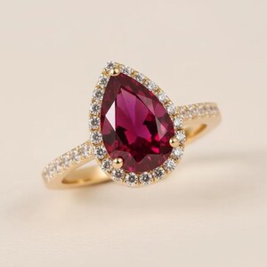 Pode incluir: Um anel de ouro com uma pedra preciosa vermelha escura em forma de pêra, rodeada por um halo de pequenas pedras transparentes. A banda do anel também é adornada com pequenas pedras transparentes. Fundo neutro.