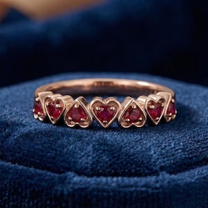 May include: A rose gold ring featuring a series of heart-shaped settings, each holding a vibrant red gemstone. The ring is set against a deep blue velvet surface, highlighting the ring's design and color.
