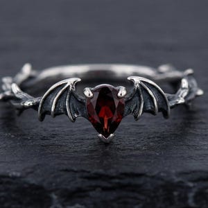 May include: A silver ring featuring a bat wing design with a pear-shaped, deep red gemstone at the center. The ring has a detailed, gothic aesthetic, with intricate detailing on the band and wings. The ring is displayed on a dark, textured surface.