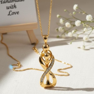 Puede incluir: Un collar con colgante dorado con un diseño único de símbolo de infinito, adornado con pequeños cristales transparentes. El collar cuelga de una delicada cadena. Un pequeño letrero dice "Handmade with Love" en el fondo.