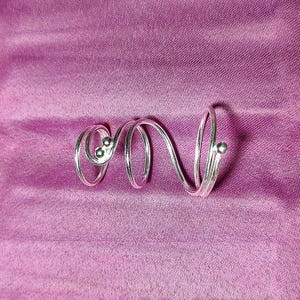 May include: A silver-colored ring with a unique, swirling design. The ring features a series of loops and curves, with small, round embellishments. The ring is set against a purple background.