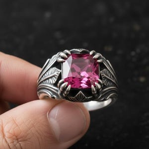 Peut inclure: Une bague en argent ornée d'une grosse pierre rose carrée. La bague présente un design orné avec des griffes retenant la pierre et des motifs de feuilles sur le bandeau. La bague est tenue par les doigts d'une personne.