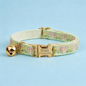 May include: A light green pet collar featuring a floral embroidery pattern in pink and gold thread. The collar includes a gold-toned metal buckle engraved with the name Silas and the number 218555789, along with a matching gold-toned jingle bell.