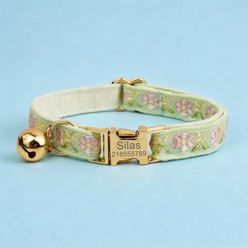 May include: A light green pet collar featuring a floral embroidery pattern in pink and gold thread. The collar includes a gold-toned metal buckle engraved with the name Silas and the number 218555789, along with a matching gold-toned jingle bell.