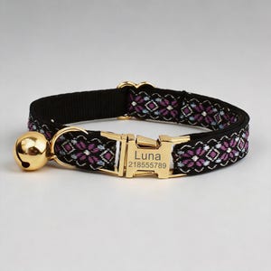 May include: A black pet collar featuring a woven floral pattern in shades of purple and light blue. The collar includes a gold-toned metal buckle engraved with the name Luna and the number 218555789, along with a matching gold-toned jingle bell.
