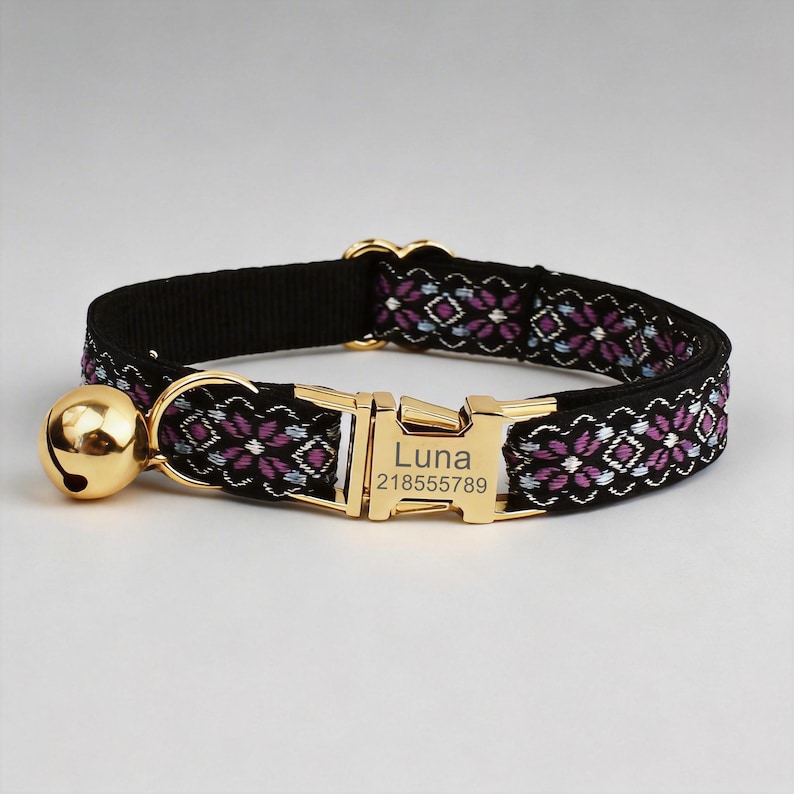 May include: A black pet collar featuring a woven floral pattern in shades of purple and light blue. The collar includes a gold-toned metal buckle engraved with the name Luna and the number 218555789, along with a matching gold-toned jingle bell.