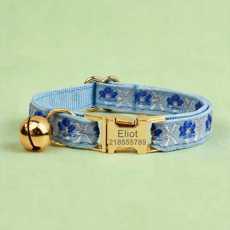 May include: A light blue pet collar featuring a repeating dark blue and silver floral pattern. The collar includes a gold-toned metal buckle engraved with the name Eliot and the number 218555789, along with a matching gold-toned metal bell.