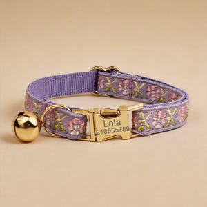 May include: A lavender pet collar featuring an embroidered floral pattern in pink and green. The collar includes a gold-toned metal buckle engraved with the name Lola and the number 218555789, along with a matching gold-toned bell attached to a D-ring.