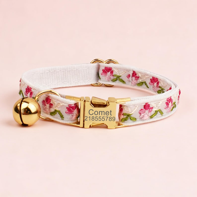 May include: A white pet collar featuring an embroidered pink floral pattern with green stems. The collar includes a gold-toned metal buckle engraved with the text Comet 218555789 and a matching gold-toned bell attached to a circular metal ring.