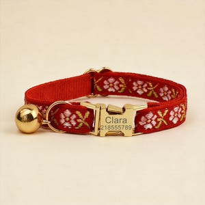 May include: A red pet collar featuring a floral embroidery pattern in pink and green. The collar includes a gold-toned metal buckle engraved with the name Clara and the number 218555789, along with a small, round, polished gold-toned bell attached.