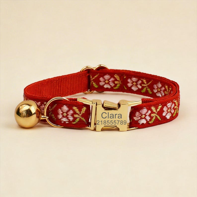 May include: A red pet collar featuring a floral embroidery pattern in pink and green. The collar includes a gold-toned metal buckle engraved with the name Clara and the number 218555789, along with a small, round, polished gold-toned bell attached.