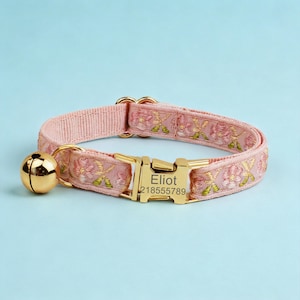 May include: A light pink pet collar featuring an embroidered floral pattern in shades of pink and green. The collar includes a gold-toned metal buckle engraved with the name Eliot and the number 218555789, along with a matching gold-toned bell.